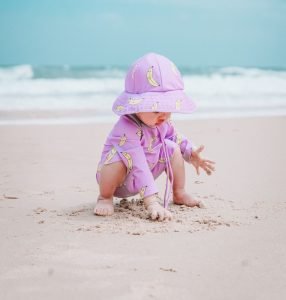 Neonato gioca sulla sabbia in spiaggia con costume e cappellino UPF 50+ a fantasia