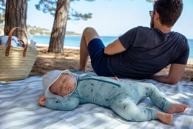 Neonato che dorme su telo da spiaggia con tutina a manica lunga e cappellino, protezione solare alternativa alla crema sotto i 6 mesi